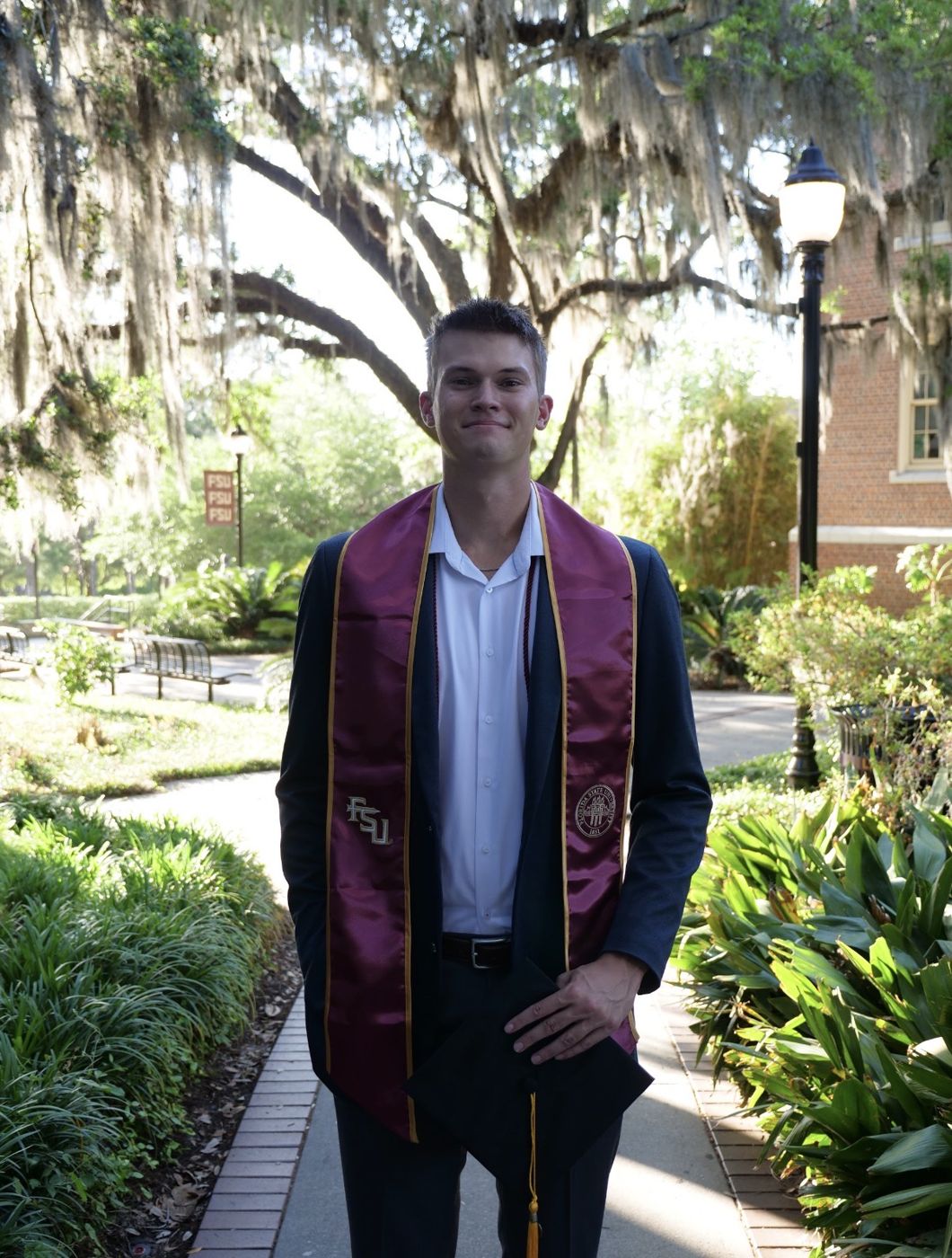 Cole Pridgen in a navy blazer and Florida State graduation stole, standing under oak trees on campus